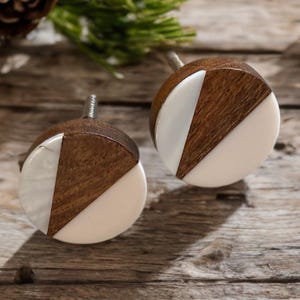 May include: Two round wooden cabinet knobs with a geometric design. Each knob features a section of dark brown wood and a section of white resin. The knobs are set against a rustic wooden background, suggesting a home decor theme.