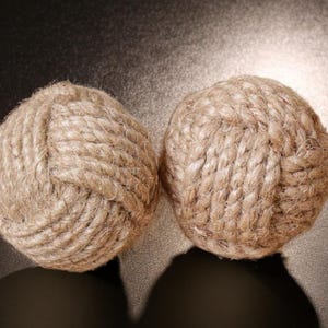 Pode incluir: Dois objetos decorativos esf&eacute;ricos feitos de corda natural bem enrolada. A corda &eacute; de cor castanho claro e as esferas est&atilde;o sobre uma superf&iacute;cie escura. Os objetos t&ecirc;m uma est&eacute;tica n&aacute;utica ou r&uacute;stica.