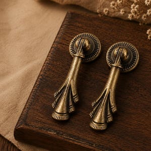 May include: Two antique brass drawer pulls with a decorative design. The pulls have a circular backplate and a detailed, elongated handle. They are set on a dark wooden surface, suggesting a vintage or classic aesthetic. The brass finish adds a touch of elegance.