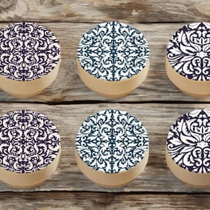 May include: Six decorative round objects with a beige base and patterned tops. The patterns include white and navy blue, and white and dark teal designs. The objects are arranged on a wooden surface.