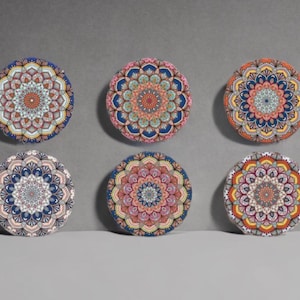 Può includere: Sei sottobicchieri rotondi con intricati disegni mandala. Ogni sottobicchiere presenta una tavolozza di colori unica, tra cui sfumature di blu, arancione, rosa e giallo. I sottobicchieri sono disposti su una superficie grigia, mostrando i loro motivi dettagliati.