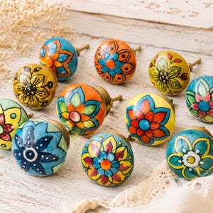 May include: A collection of colorful, hand-painted ceramic cabinet knobs. Each knob features a unique floral design in shades of blue, orange, yellow, and green. The knobs have brass hardware and are arranged on a white surface.