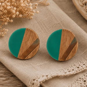 May include: A pair of round stud earrings. Each earring features a teal green resin half and a wooden half with diagonal wood grain and a brown stripe. The earrings are displayed on a beige linen surface.