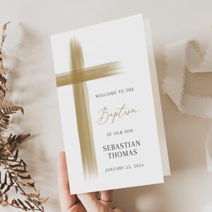 May include: A folded baptism invitation with a gold cross design and the text "Welcome to the Baptism of our Son Sebastian Thomas January 22, 2024." A hand holds the invitation. Dried fern and white ribbon are also in the image.