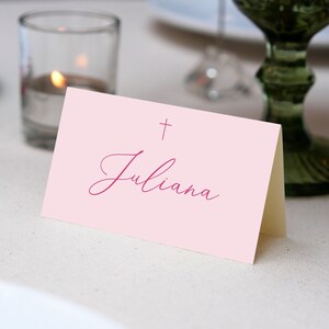 May include: A pink place card with a small cross and the name "Juliana" written in cursive in a dark pink color.