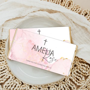 Puede incluir: Dos envoltorios de barra de chocolate rosa y dorado con una cruz y el texto "Amelia Rose" y "Gracias por celebrar con nosotros".