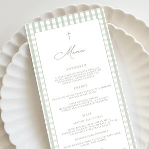May include: A menu card with a light green gingham border and a white interior. The menu features a cross at the top, the word "Menu" in cursive, and a list of dishes including antipasto, entree, and main courses. The card is on a white plate.