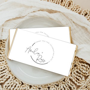 Puede incluir: Tarjeta blanca con borde dorado, con un diseño circular simple con el texto "Gracias por celebrar con nosotros" y los nombres "Amelia + Ross" en cursiva.
