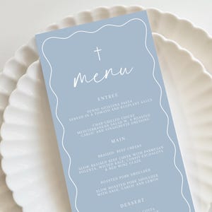 Pode incluir: Um cartão de menu azul claro com uma cruz branca e a palavra "Menu" escrita em itálico. O menu lista entradas, pratos principais e sobremesas. As entradas incluem penne siciliana e frango grelhado. Os pratos principais incluem bochechas de carne bovina braseadas e ombro de porco assado. As sobremesas incluem uma sobremesa chamada "Cotta".