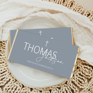 Op de afbeelding: Twee grijze chocoladeverpakkingen met gouden rand. De bovenste verpakking heeft een wit kruis en de tekst "THOMAS". De onderste verpakking heeft een wit kruis, de tekst "THOMAS" en de cursieve tekst "Jackson" gevolgd door "THANK YOU FOR CELEBRATING WITH US".