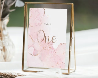 Op de afbeelding: Een tafelnummerkaart met een gouden frame en een roze en gouden aquarelontwerp. De kaart heeft de tekst "Table One" in een bruin lettertype.