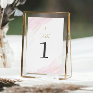 May include: A table number card in a gold-colored frame. The card features a watercolor design with pink and gold accents. The word "Table" and the number "1" are displayed in elegant script. A gold cross is at the top.