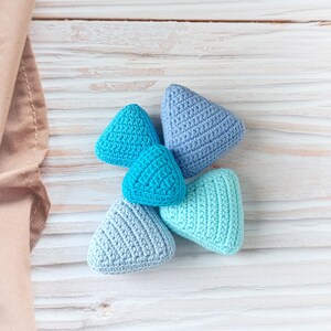 Puede incluir: Una colección de cinco objetos triangulares de ganchillo en tonos azules y turquesas. Los artículos hechos a mano están dispuestos sobre una superficie clara con vetas de madera, con una tela de color neutro en el fondo.