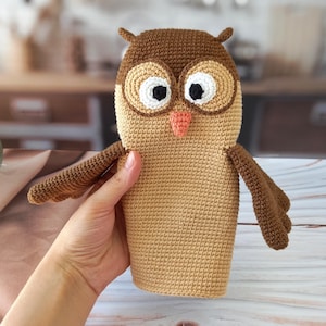 May include: Crocheted owl in shades of brown and tan. The owl has large, round eyes with black pupils and a peach-colored beak. The wings are outstretched, and the owl is held by a hand. The owl is a handmade item.