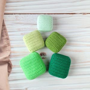 May include: Five square crochet items in shades of green, arranged on a light wood surface. The colors range from light green to dark green. The items are handmade and have a textured appearance.