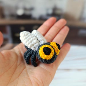 Puede incluir: Una abeja de ganchillo hecha a mano con alas blancas, una cabeza negra y amarilla y un cuerpo gris oscuro. La abeja es un pequeño artículo artesanal detallado, probablemente para decoración o como juguete. La abeja se sostiene en la mano de una persona.