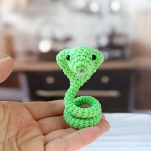 Puede incluir: Una pequeña serpiente de ganchillo hecha a mano en verde brillante. La serpiente está enrollada con una capucha acampanada y ojos de cuentas negras. El artículo artesanal se sostiene en una mano, con un fondo borroso.