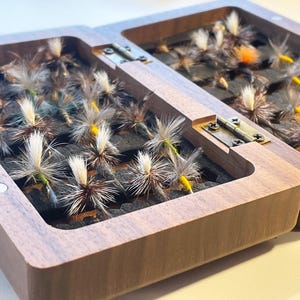 Może przedstawiać: Otwarta drewniana skrzynka do łowienia much z pokrywą na zawiasach, wypełniona różnymi sztucznymi muchami. Muchy mają białe, brązowe i żółte akcenty i są ułożone na ciemnej powierzchni. Pudełko wykonane jest z jasnobrązowego drewna.