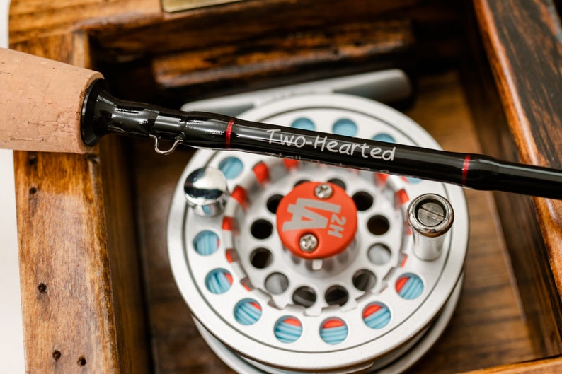 May include: A black fly fishing rod with a cork handle and the inscription "Two-Hearted" on the rod. The rod is paired with a silver fly reel with a red and blue spool.