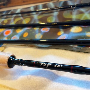 May include: A black fly fishing rod with a brown cork handle and a red line. The rod is labeled "7.5 ft 2wt".