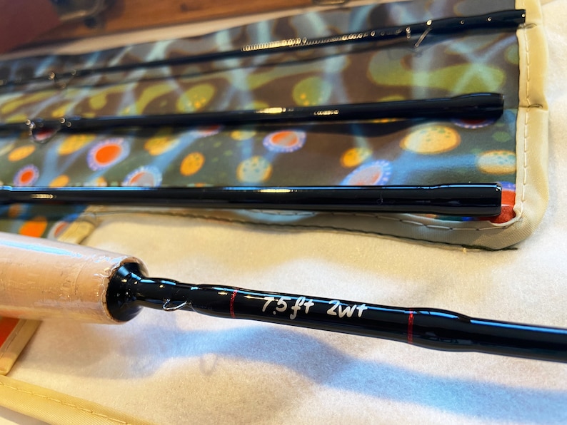 May include: A black fly fishing rod with a brown cork handle and a red line. The rod is labeled "7.5 ft 2wt".