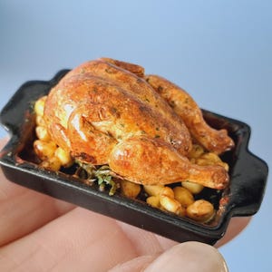 Puede incluir: Pollo asado en miniatura con piel dorada, servido en una fuente negra con patatas y hierbas. El pequeño alimento es una réplica detallada, ideal para casas de muñecas o exhibiciones en miniatura.