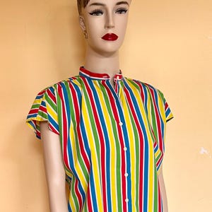 May include: A mannequin wearing a colorful striped shirt with red, yellow, green, and blue vertical stripes. The shirt has a mandarin collar and short sleeves. The mannequin is also wearing black shorts with a red and white pattern.