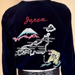 Può includere: Un giubbotto bomber in velluto nero con una mappa del Giappone ricamata sul retro. Il design include la parola Japan in corsivo rosso, una raffigurazione del monte Fuji, una tigre e vari nomi di città tra cui Tokyo, Osaka e Kyoto.