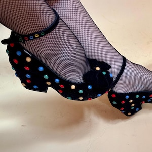 May include: Black velvet platform shoes with colorful polka dots and ankle straps. The shoes have a bow detail on the toe and a low heel. The shoes are worn with black fishnet stockings.