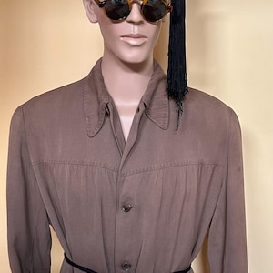 May include: A maroon fez hat with white beaded text reading LUXOR DIRECTOR and a central emblem, worn by a mannequin with round tortoiseshell sunglasses. The mannequin wears a long-sleeved brown button-down shirt with a black tie-waist belt.