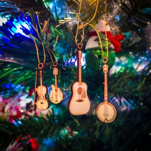 May include: Four wooden ornaments shaped like musical instruments hang from a Christmas tree. The ornaments are a guitar, a violin, a banjo, and a mandolin. The ornaments are all painted in a natural wood color.