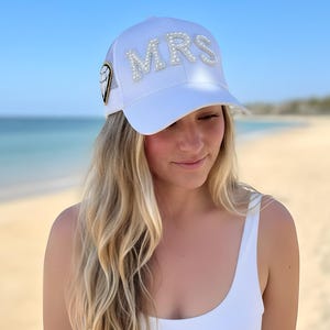 May include: A white baseball cap with the word "MRS" spelled out in pearl-like embellishments. The cap is worn by a person wearing a white swimsuit on a beach with blue water and sky in the background.