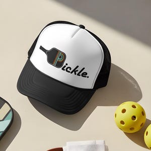 May include: A white and black trucker hat featuring a graphic of a pickleball paddle with rainbow stripes and the text pickle. The set includes a geometric patterned paddle, two yellow perforated pickleballs, and a cream-colored canvas bag.