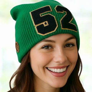 May include: A green knit beanie with the number "52" in black with gold trim. The beanie has a ribbed texture and a small brown circular patch on the side. The person in the image is smiling.