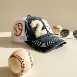 May include: A navy blue baseball cap with the number 25 in white and gold, a white mesh back, and a baseball patch. Two baseballs and a pair of sunglasses are also in the image.