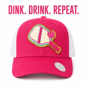 May include: A hot pink baseball cap with white mesh back panels. The front features a patch of a pickleball paddle and ball with gold glitter trim. The text DINK. DRINK. REPEAT. is printed in pink above the cap on a white background.