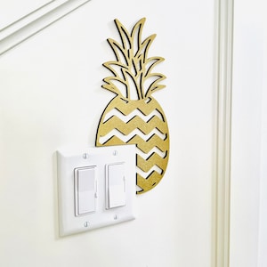 May include: A gold pineapple-shaped wall decor with a chevron pattern on the body of the pineapple. The decor is mounted on a white wall next to a white light switch.