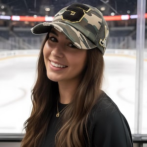 May include: A camouflage print baseball cap with a distressed brim, featuring a large black number 5 patch with gold trim on the front and a gold-colored hockey stick emblem on the side. The cap has a mesh back panel and a structured crown.