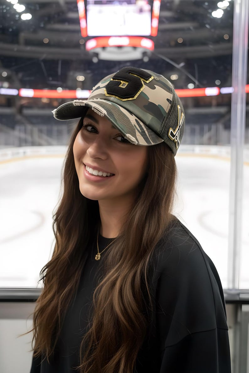 May include: A camouflage print baseball cap with a distressed brim, featuring a large black number 5 patch with gold trim on the front and a gold-colored hockey stick emblem on the side. The cap has a mesh back panel and a structured crown.