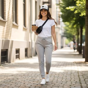 May include: A white t-shirt, light gray drawstring sweatpants, white sneakers, a black crossbody bag, and a white and black baseball cap with the word pickle printed on the front. The outfit is completed with a brown disposable coffee cup.