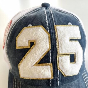 May include: A denim baseball cap with a white mesh back. The front displays the number "25" in white with gold glitter trim. White stitching details the cap's design.