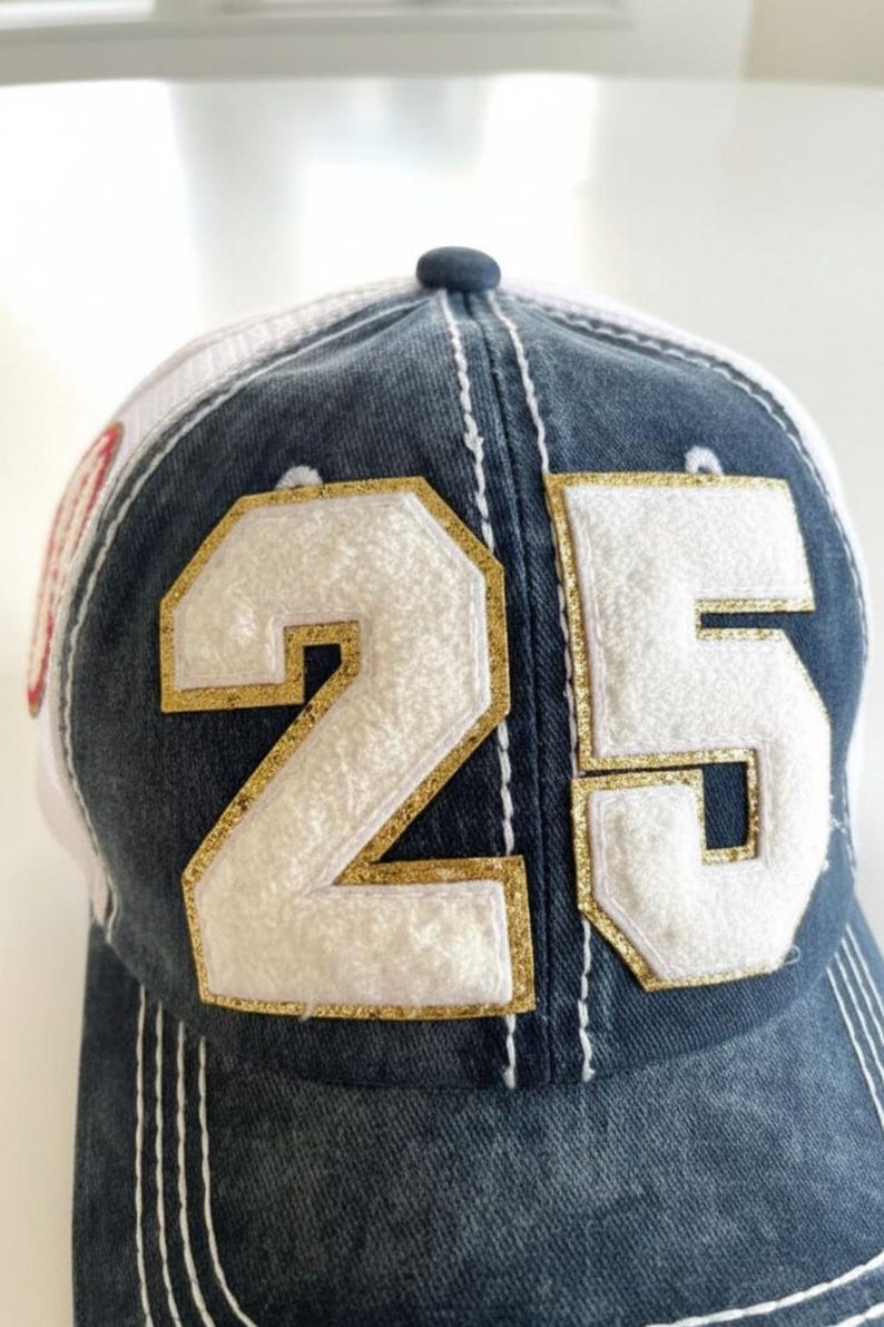 May include: A denim baseball cap with a white mesh back. The front displays the number "25" in white with gold glitter trim. White stitching details the cap's design.