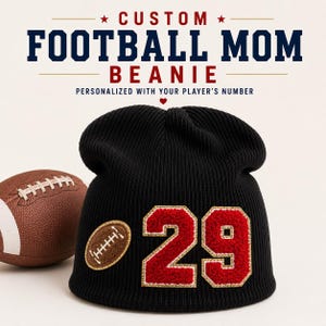 May include: A black ribbed knit beanie featuring a red and gold patch with the number 29 and a small brown football-shaped patch. Above the beanie, text reads CUSTOM FOOTBALL MOM BEANIE PERSONALIZED WITH YOUR PLAYER'S NUMBER. A brown football is visible.