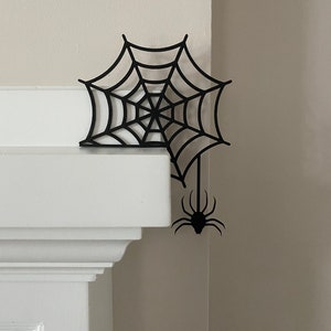 May include: Black spider web and spider cutout decoration for Halloween. The web is attached to a corner of a white wall.
