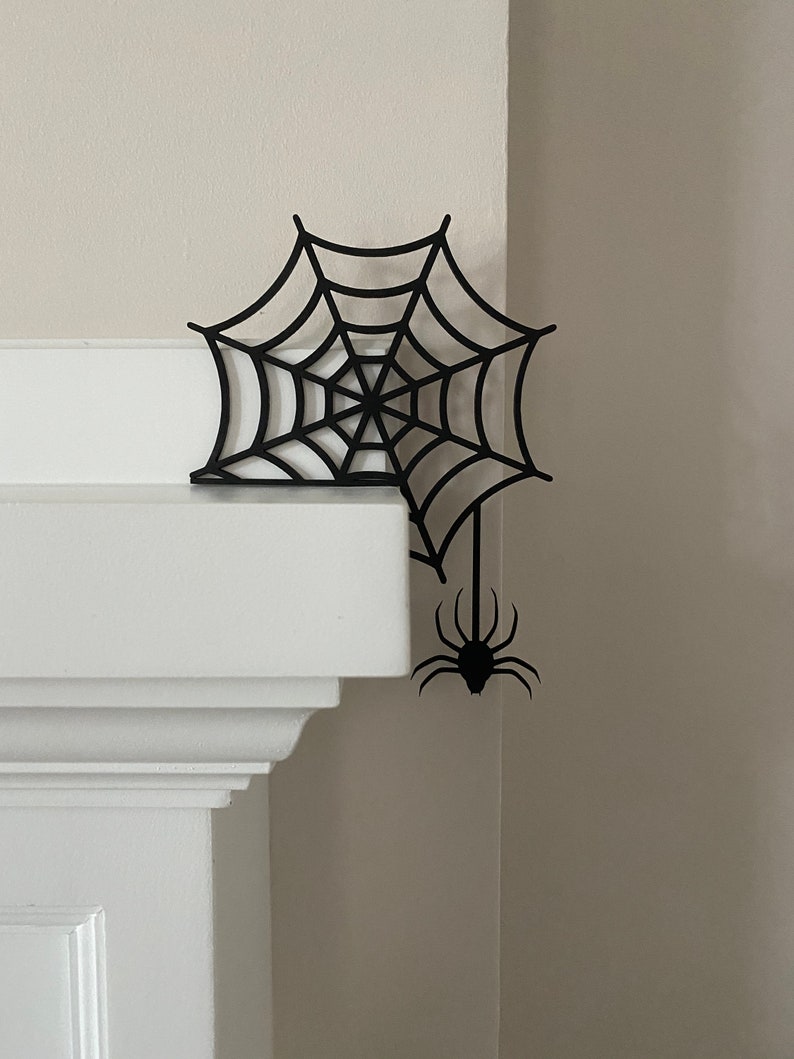 May include: Black spider web and spider cutout decoration for Halloween. The web is attached to a corner of a white wall.
