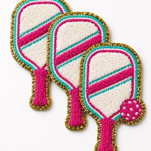 Peut inclure: Trois &eacute;cussons brod&eacute;s en forme de raquettes de pickleball. Chaque raquette pr&eacute;sente un centre textur&eacute; blanc, une bande diagonale rose vif, une bordure turquoise et un contour &agrave; paillettes dor&eacute;es. Un &eacute;cusson inclut une petite balle rose &agrave; pois blancs.