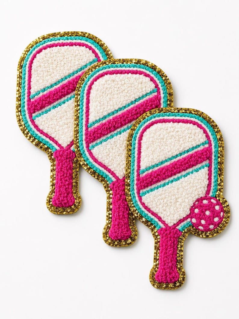 Peut inclure: Trois &eacute;cussons brod&eacute;s en forme de raquettes de pickleball. Chaque raquette pr&eacute;sente un centre textur&eacute; blanc, une bande diagonale rose vif, une bordure turquoise et un contour &agrave; paillettes dor&eacute;es. Un &eacute;cusson inclut une petite balle rose &agrave; pois blancs.