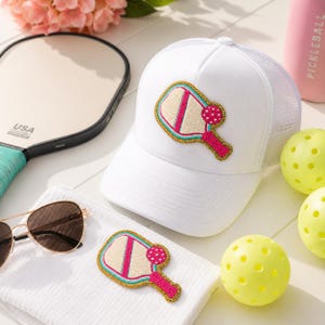 Peut inclure: Une casquette de camionneur blanche orn&eacute;e d'un &eacute;cusson brod&eacute; repr&eacute;sentant une raquette et une balle de pickleball en rose, beige et bleu sarcelle avec une bordure dor&eacute;e. La casquette poss&egrave;de un panneau avant solide et un dos en filet.