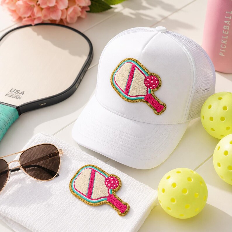 Peut inclure: Une casquette de camionneur blanche orn&eacute;e d'un &eacute;cusson brod&eacute; repr&eacute;sentant une raquette et une balle de pickleball en rose, beige et bleu sarcelle avec une bordure dor&eacute;e. La casquette poss&egrave;de un panneau avant solide et un dos en filet.