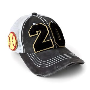 May include: A black baseball cap with a distressed finish, featuring the number 20 in black felt with gold trim on the front panels. The cap has a white mesh back, white stitching on the brim, and a circular yellow softball patch on the side.